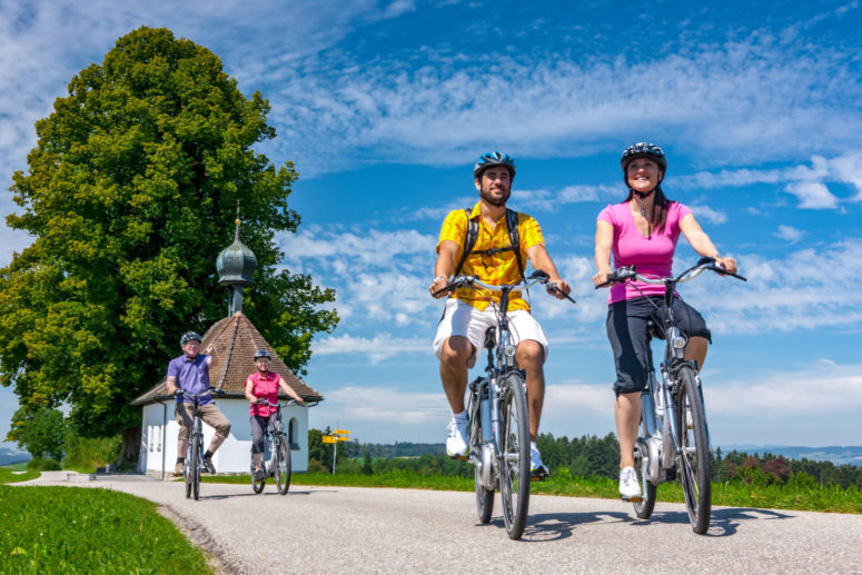 Vier Personen genießen Bayreuth für Aktive, als sie an einem sonnigen Tag mit dem Fahrrad einen Feldweg entlang fahren, vorbei an einer kleinen weißen Kapelle und einem großen Baum, umgeben von blauem Himmel und grünen Feldern. Alle tragen Helme und bunte, legere Kleidung.