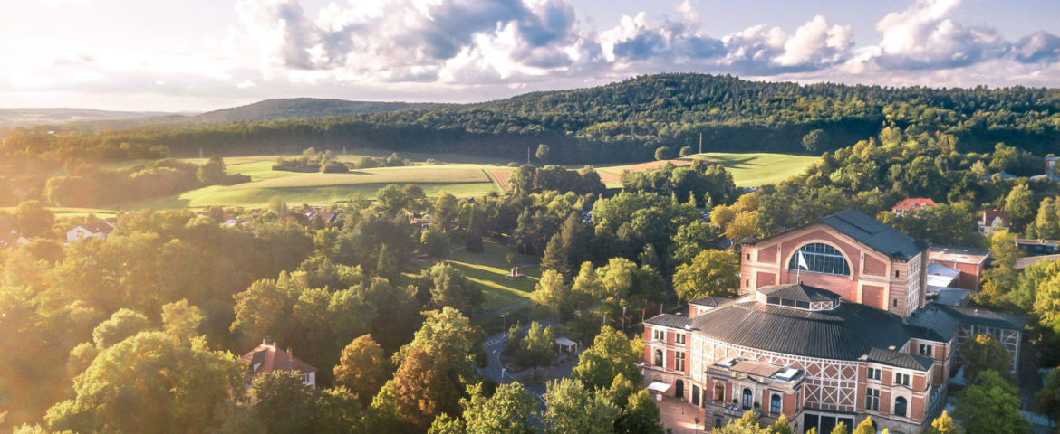 Luftaufnahme des Gebäudes der Bayreuther Festspiele, umgeben von grünen Bäumen, mit sanften Hügeln und offenen Feldern im Hintergrund unter einem teilweise bewölkten Himmel bei Sonnenuntergang.