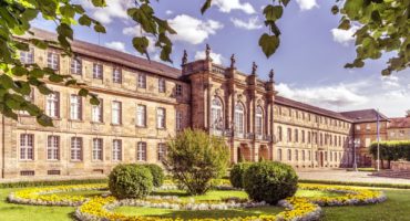 Ein prächtiges historisches Gebäude mit Statuen auf dem Dach steht hinter einem gepflegten Garten mit runden Blumenbeeten, gestutzten Büschen und leuchtend gelben und weißen Blumen unter einem strahlend blauen Himmel.