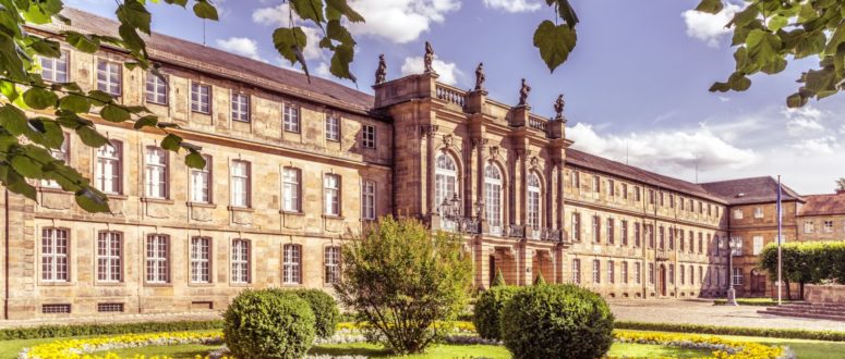 Ein prächtiges historisches Gebäude mit Statuen auf dem Dach steht hinter einem gepflegten Garten mit runden Blumenbeeten, gestutzten Büschen und leuchtend gelben und weißen Blumen unter einem strahlend blauen Himmel.