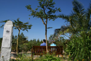 Zwei Personen sitzen auf einer Holzbank in einem üppigen, grünen Garten unter einem strahlend blauen Himmel, umgeben von Bäumen und Pflanzen. Links neben der Bank steht ein großes Steinschild mit Text.