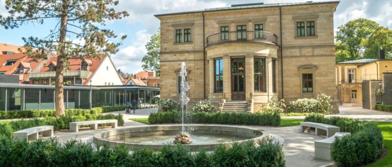 Ein Steingebäude mit großen Fenstern, einer zentralen Treppe und einem runden Springbrunnen davor, umgeben von Bänken, grünem Rasen und Bäumen an einem sonnigen Tag - perfekt für beeindruckende Pressefotos.