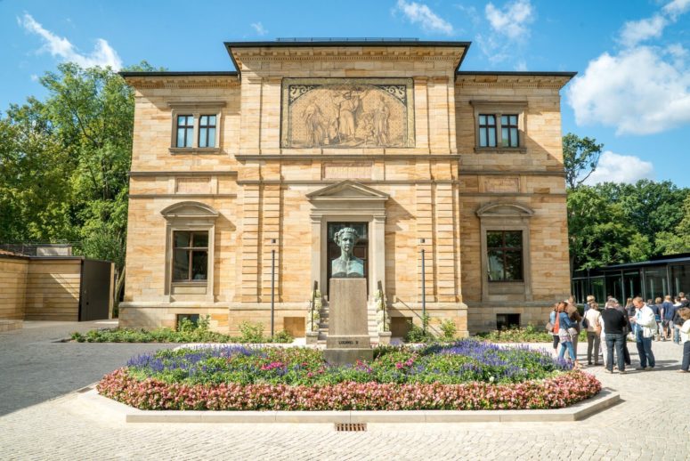 Ein historisches Steingebäude mit einer Statue davor, umgeben von bunten Blumenbeeten. Mehrere Menschen stehen und gehen in der Nähe an einem sonnigen Tag mit blauem Himmel und vereinzelten Wolken.