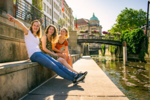Drei junge Frauen sitzen auf einem sonnigen Kanalufer in der Stadt, lächeln und lachen während ihrer Sightseeing-Tour. Eine zeigt aufgeregt auf etwas in der Ferne, mit einer Brücke, Wasser und historischen Gebäuden im Hintergrund.