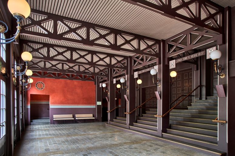 Eine geräumige Halle mit dunkelroten und braunen Balken, kugelförmigen Wandlampen, einer Bank, einer roten Wand mit einer Uhr und einer Treppe, die nach oben führt. Der Boden ist aus Stein und der Raum wird durch natürliches Licht hell erleuchtet.