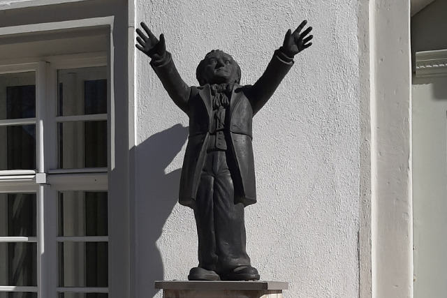 Eine dunkle Statue einer Person in einem langen Mantel steht mit erhobenen Armen vor einem hellen Gebäude mit Fenstern und architektonischen Details.