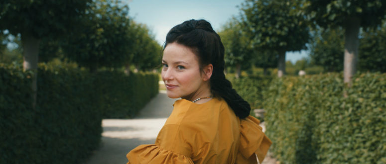 Eine Frau in einem senfgelben Kleid steht auf einem von gepflegtem Grün gesäumten Kiesweg und blickt mit einem leichten Lächeln über ihre Schulter in Richtung Kamera. Die Szene erinnert an die elegante Drehorte Die Kaiserin Netflix unter einem klaren blauen Himmel.