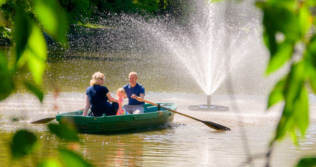Eine dreiköpfige Familie genießt einen sonnigen Tag und rudert ein grünes Boot auf einem Teich in der Nähe eines großen Springbrunnens, umgeben von üppig grünen Bäumen und Blättern.