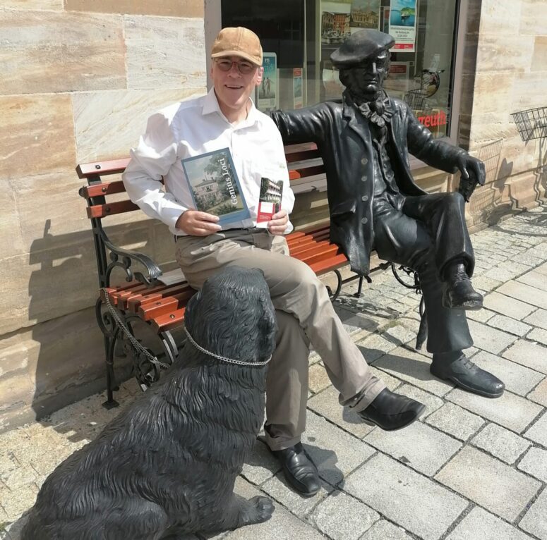 Der Autor Dr. Frank Piontek sitzt auf einer Holzbank vor der Tourist Information in Bayreuth, neben ihm sitzt eine Plastik, die den Komponisten Richard Wagner darstellt, mit auf der Bank, in den Händen hält der Autor sein neues Buch und einen Flyer