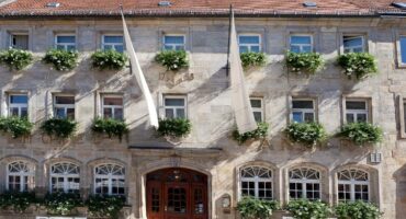 Historisches Steingebäude mit gewölbter Holztür, großen Fenstern mit weißen Rahmen und grünen Blumenkästen, braunen Fahnen über dem Eingang und weißen Bänken davor.