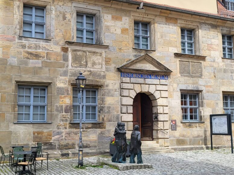 Das Historische Museum befindet sich in einem Steingebäude mit zahlreichen blau umrahmten Fenstern und verfügt über lebensgroße Bronzestatuen am Eingang. Ein altmodischer Laternenpfahl und Sitzgelegenheiten im Freien zieren das Kopfsteinpflaster im Außenbereich.