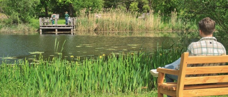 Ein Mann sitzt auf einer Holzbank am Ufer eines Teiches und blickt aufs Wasser. Im Hintergrund stehen zwei Menschen auf einem kleinen Steg, der von Schilf und grünen Bäumen umgeben ist.