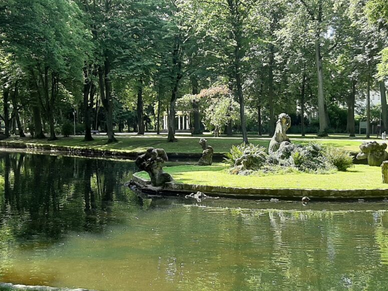 Eine friedliche Parkszene mit einem Teich, einer kleinen Insel mit Steinskulpturen und üppig grünen Bäumen. Das Sonnenlicht fällt durch die Blätter und wirft Schatten auf das Gras und das Wasser.