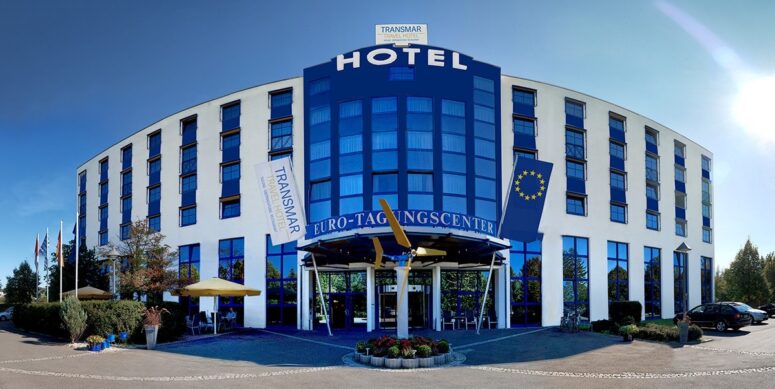 Ein modernes, geschwungenes weiß-blaues Hotelgebäude mit vielen Fenstern, beschriftet mit HOTEL und EURO-TAGUNGSCENTER, mit Fahnen und Bäumen davor und einem Kreisverkehr, der zum Eingang führt.