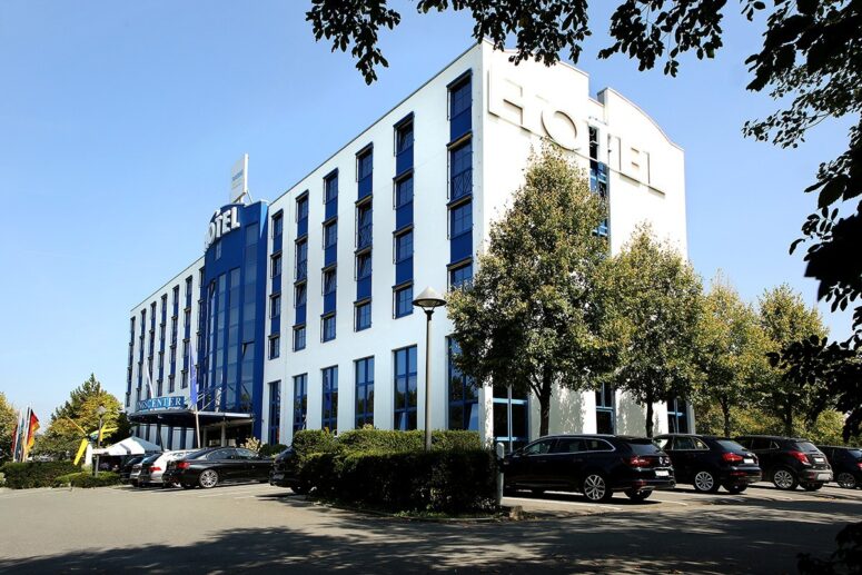 Ein modernes weißes Hotelgebäude mit großen blau getönten Fenstern, mehreren Stockwerken und HOTEL-Beschriftung. Vor dem Gebäude sind mehrere Autos geparkt, und Bäume säumen den Parkplatz unter dem klaren Himmel.