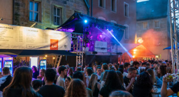 Eine Menschenmenge genießt ein Abendkonzert auf dem Bayreuther Bürgerfest auf einem Stadtplatz mit bunten Bühnenlichtern, einem erhöhten DJ-Pult und Menschen, die sich in der Nähe von Getränkeständen unter Regenschirmen aufhalten. Im Hintergrund sind historische Gebäude zu sehen, die sich perfekt für Bilder eignen.