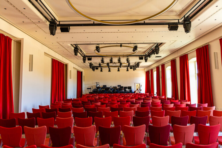In einem hell erleuchteten Saal mit roten Vorhängen und großen Fenstern stehen Reihen leerer roter Stühle vor einer Bühne mit Musikinstrumenten und Scheinwerfern.
