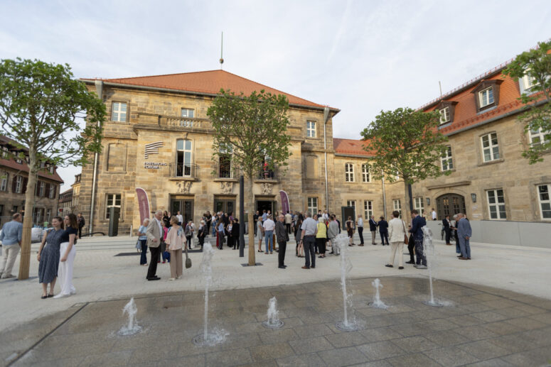 Die Menschen versammeln sich auf einem Innenhof mit Springbrunnen vor einem historischen Steingebäude mit roten Dächern und Bäumen. Einige der Anwesenden unterhalten sich, andere gehen umher, und am Eingang sind Transparente zu sehen.