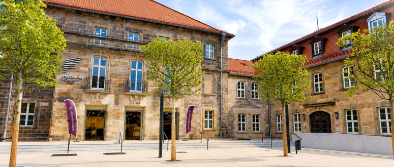 Ein historisches Steingebäude mit roten Dachziegeln, hohen Fenstern und einem Innenhof, der von jungen, grünen Bäumen gesäumt ist, unter einem blauen Himmel mit vereinzelten Wolken. Neben dem Eingang wehen zwei violette Fahnen.