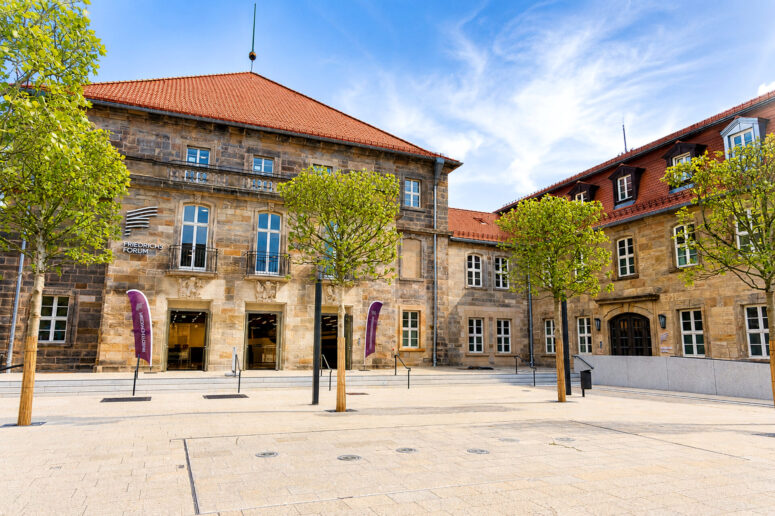 Ein historisches Steingebäude mit roten Dachziegeln, hohen Fenstern und einem Innenhof, der von jungen, grünen Bäumen gesäumt ist, unter einem blauen Himmel mit vereinzelten Wolken. Neben dem Eingang wehen zwei violette Fahnen.
