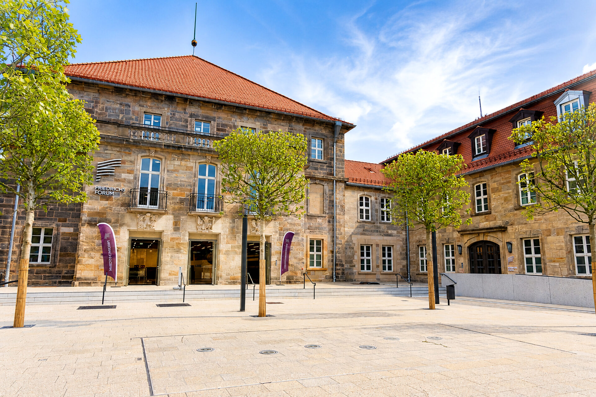Ein historisches Steingebäude mit roten Dachziegeln, hohen Fenstern und einem Innenhof, der von jungen, grünen Bäumen gesäumt ist, unter einem blauen Himmel mit vereinzelten Wolken. Neben dem Eingang wehen zwei violette Fahnen.
