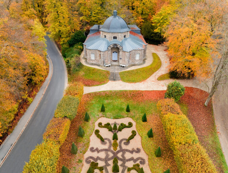 Luftaufnahme eines kleinen, historischen Steingebäudes mit Kuppeldach, eingebettet zwischen leuchtenden Herbstbäumen und einem gepflegten Garten mit geometrischen Mustern, neben einer gewundenen Straße in einem der malerischen Parks von Bayreuth.