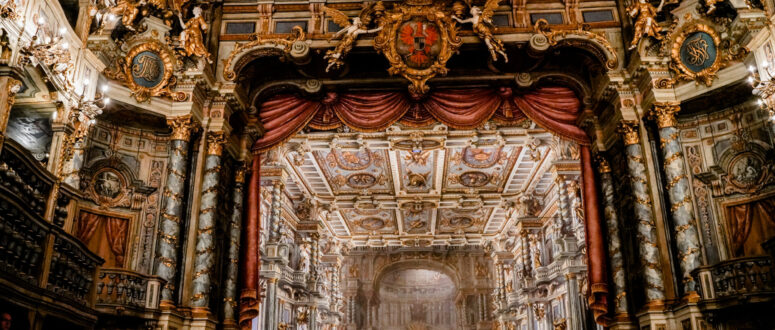 Opulenter Opernhaus-Innenraum mit üppig verzierter Barockdecke, goldenen Akzenten, funkelnden Kronleuchtern, roten Vorhängen und aufwändigen architektonischen Details, alles von den Zuschauerplätzen aus gesehen in Richtung der großen Bühne.