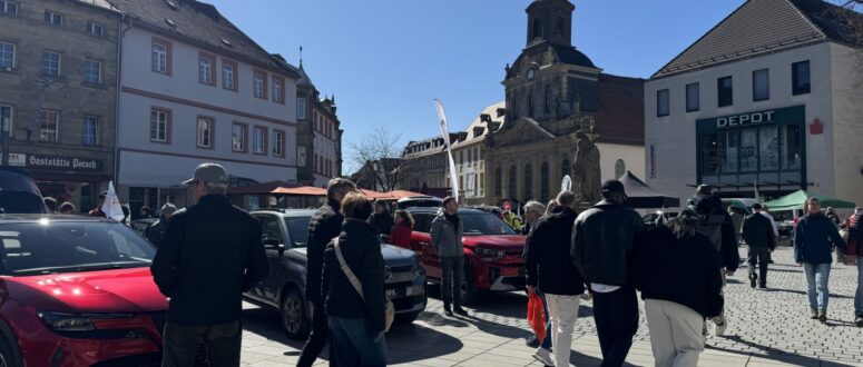Menschen gehen während der Mobilitätstage über einen sonnigen Stadtplatz, auf dem Autos geparkt sind. Historische und moderne Gebäude, darunter eine Kirche und das DEPOT-Geschäft, säumen den Hintergrund. Einige Zelte und Marktstände sind zu sehen.