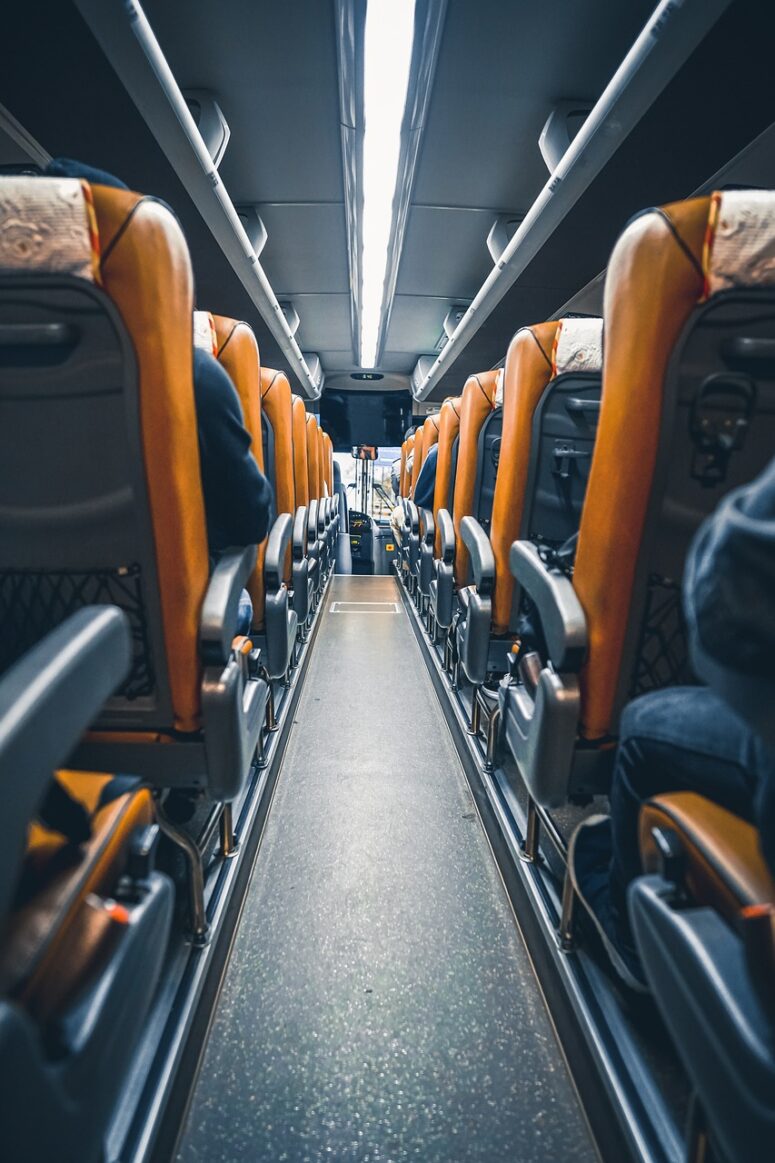 Blick auf den Gang im Inneren eines modernen Busses mit orangefarbenen und grauen Sitzen auf beiden Seiten, Gruppen von Fahrgästen sitzen und schauen nach vorne. Die Beleuchtung ist hell und der Bus wirkt sauber und gut gepflegt.