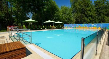 Das Freibad ist umgeben von grünen Bäumen, Liegestühlen mit grünen Sonnenschirmen und einer bunten Wandmalerei an der Rückwand an einem sonnigen Tag. Der Bereich ist sauber und einladend, mit klarem, blauem Wasser.