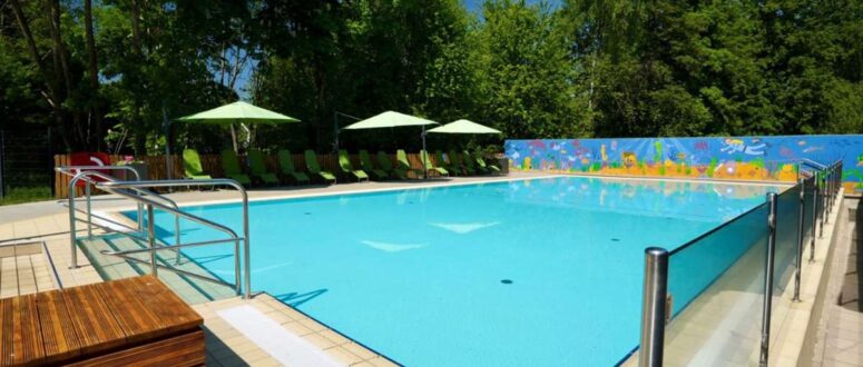 Das Freibad ist umgeben von grünen Bäumen, Liegestühlen mit grünen Sonnenschirmen und einer bunten Wandmalerei an der Rückwand an einem sonnigen Tag. Der Bereich ist sauber und einladend, mit klarem, blauem Wasser.