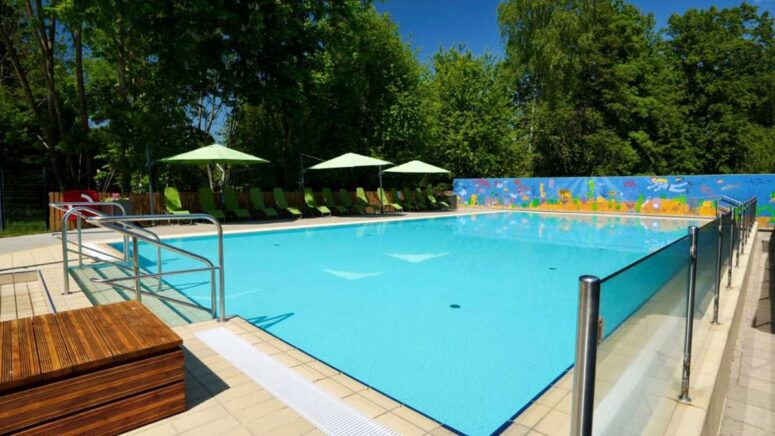 Das Freibad ist umgeben von grünen Bäumen, Liegestühlen mit grünen Sonnenschirmen und einer bunten Wandmalerei an der Rückwand an einem sonnigen Tag. Der Bereich ist sauber und einladend, mit klarem, blauem Wasser.