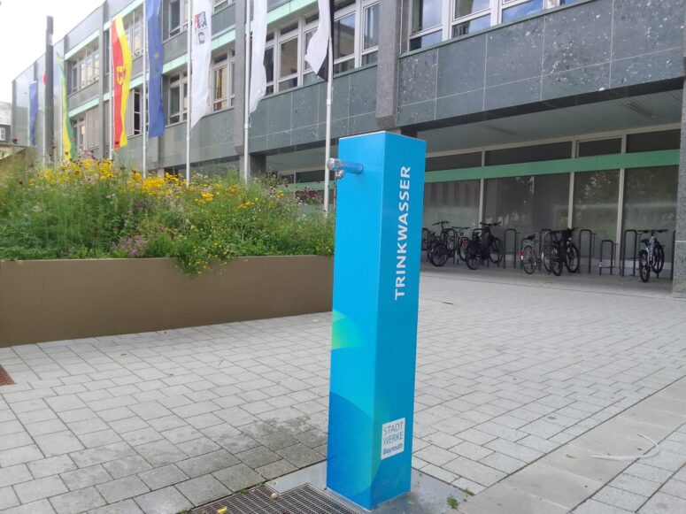 Ein blauer öffentlicher Trinkwasserbrunnen mit der Aufschrift Trinkwasser steht auf einer gepflasterten Fläche vor einem Gebäude mit Fahnen und Fahrradständern, während in einem Hochbeet in der Nähe Blumen blühen.