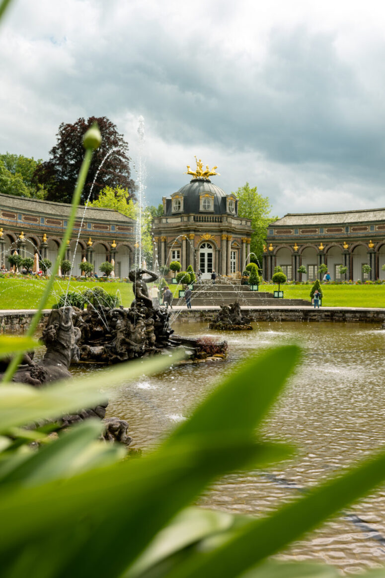 Eleganter Barockpalast mit Kuppeldach und Goldstatue, umgeben von einer geschwungenen Kolonnade und Gärten. Ein dekorativer Springbrunnen spritzt Wasser in einen großen Teich mit grünen Blättern im Vordergrund. Menschen spazieren in der Nähe unter einem bewölkten Himmel.