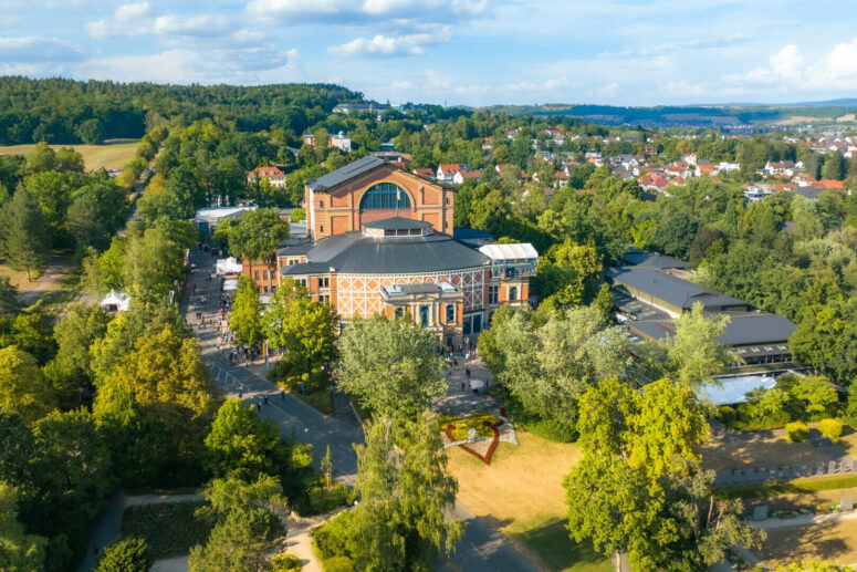 Luftaufnahme des großen, historischen Bayreuther Theaters, umgeben von üppigen grünen Bäumen, mit einer Stadt und Hügeln im Hintergrund unter einem blauen Himmel mit vereinzelten Wolken.