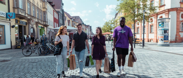 Vier junge Erwachsene gehen an einem sonnigen Tag mit Einkaufstüten auf einer Kopfsteinpflasterstraße spazieren, lächeln und unterhalten sich, während im Hintergrund Fahrräder und bunte Gebäude zu sehen sind.