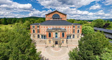 Ein großes, historisches Theater mit einer Fassade aus Ziegeln und Stein, umgeben von grünen Bäumen unter einem blauen Himmel mit vereinzelten Wolken. Das Gebäude hat Rundbogenfenster und steht auf einem runden Platz.