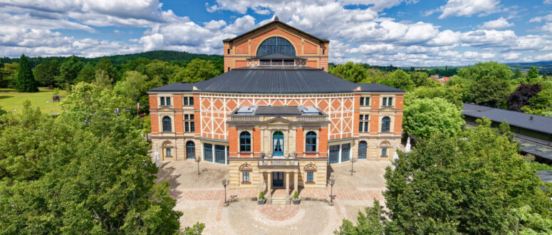 Ein großes, historisches Theater mit einer Fassade aus Ziegeln und Stein, umgeben von grünen Bäumen unter einem blauen Himmel mit vereinzelten Wolken. Das Gebäude hat Rundbogenfenster und steht auf einem runden Platz.
