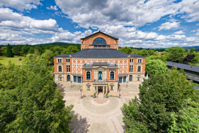 Ein großes, historisches Theater mit einer Fassade aus Ziegeln und Stein, umgeben von grünen Bäumen unter einem blauen Himmel mit vereinzelten Wolken. Das Gebäude hat Rundbogenfenster und steht auf einem runden Platz.