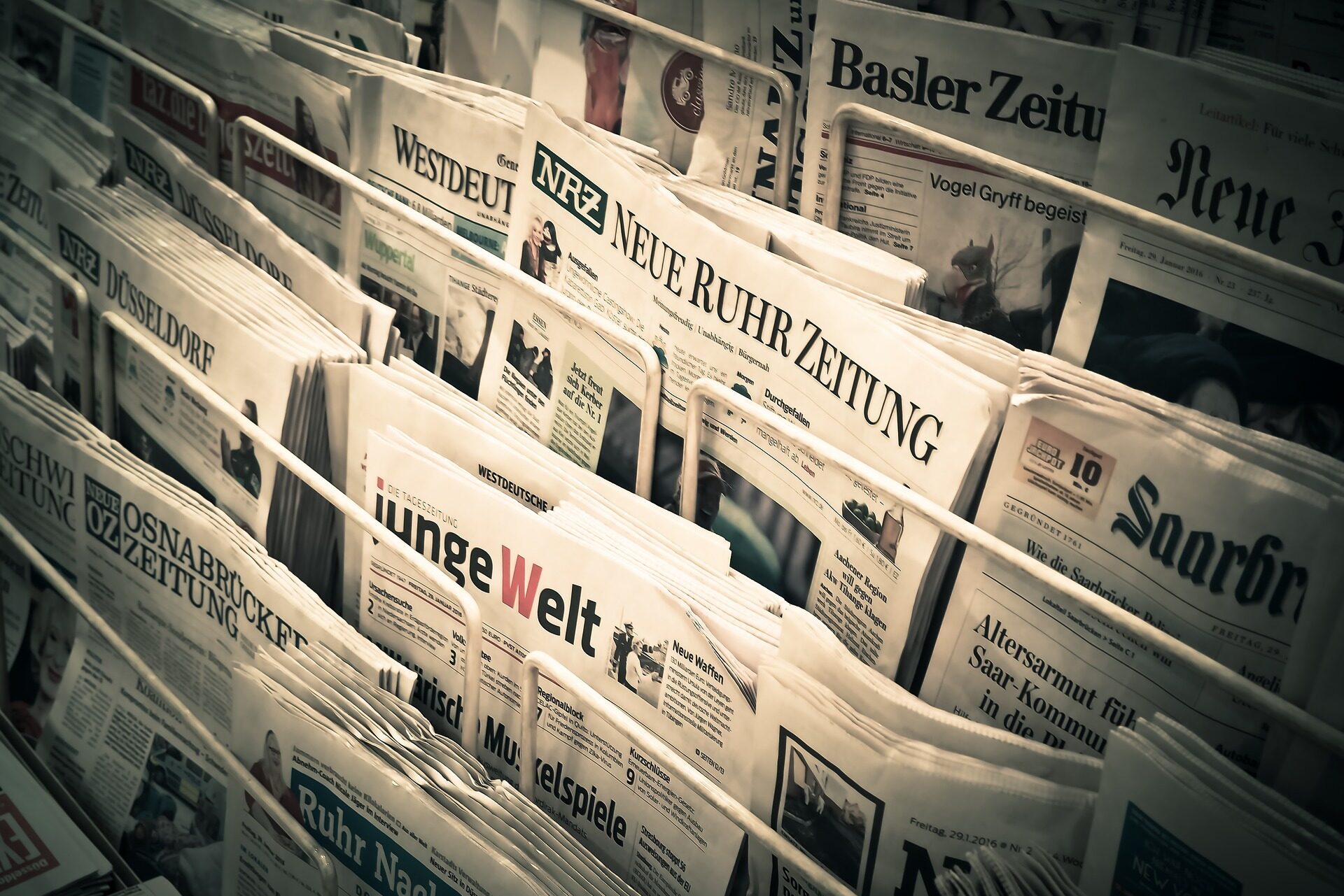 Ein Regal, gefüllt mit verschiedenen deutschsprachigen Zeitungen, darunter Titel wie "Neue Ruhr Zeitung", "Junge Welt" und "Basler Zeitung", ordentlich in überlappenden Reihen angeordnet.