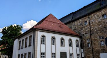 Ein weißes, zweistöckiges Gebäude mit großen Bogenfenstern und einem roten Ziegeldach steht neben einem Steinhaus unter einem blauen Himmel mit Schäfchenwolken, die Spuren des jüdischen Lebens widerspiegeln. Die Straße davor ist leer und wird von einem Fahrradweg gesäumt.