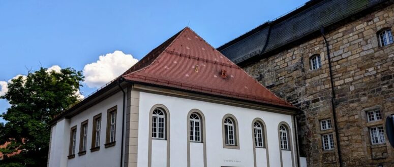Ein weißes, zweistöckiges Gebäude mit großen Bogenfenstern und einem roten Ziegeldach steht neben einem Steinhaus unter einem blauen Himmel mit Schäfchenwolken, die Spuren des jüdischen Lebens widerspiegeln. Die Straße davor ist leer und wird von einem Fahrradweg gesäumt.