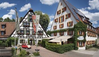 Zwei traditionelle Fachwerkhäuser mit steilen, dreieckigen Dächern und verschlossenen Fenstern, eines davon mit Efeu bewachsen. Ein Innenhof mit einem roten Oldtimer und Fahnen davor, unter einem blauen Himmel mit vereinzelten Wolken.