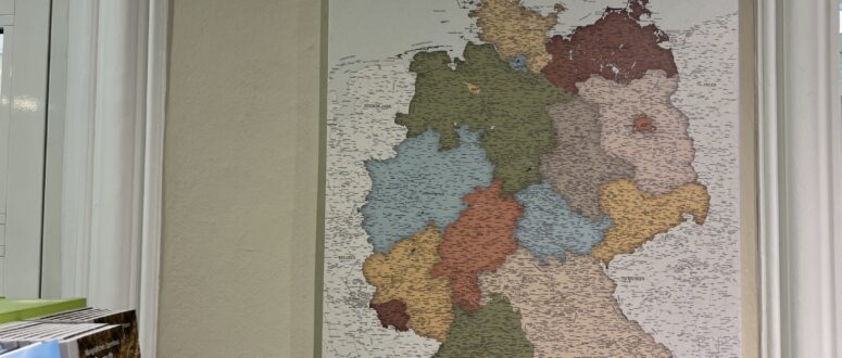 Eine detaillierte Deutschlandkarte mit den Bundesländern in verschiedenen Farben hängt an einer beigen Wand neben einem Fenster, über einem grünen Regal mit Broschüren der Tourist-Information, das in der unteren linken Ecke teilweise sichtbar ist.