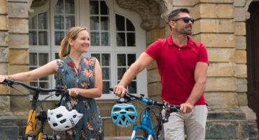 Eine lächelnde Frau in einem geblümten Kleid und ein Mann in einem roten Poloshirt stehen mit ihren E-Bikes auf einer Kopfsteinpflasterstraße vor einem historischen Steingebäude mit großen Fenstern und verzierten Details.