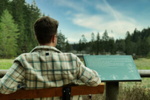 Eine Person in einer karierten Jacke sitzt auf einer Bank und liest ein Hinweisschild mit Blick auf eine grüne, bewaldete Landschaft unter einem teilweise bewölkten Himmel.