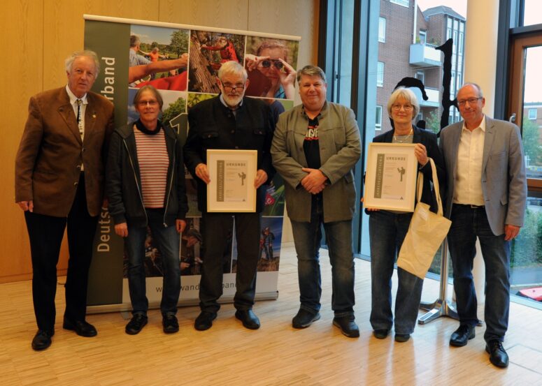 Sechs Erwachsene stehen in einem Raum, einige halten gerahmte Urkunden und eine Tragetasche in der Hand und posieren für ein Gruppenfoto vor einem Banner und einer holzgetäfelten Wand. Sie scheinen an einer offiziellen Veranstaltung oder Preisverleihung teilzunehmen.
