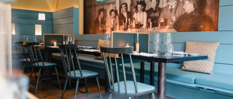Bistro „oma & enkel“ mit hellblauen Wänden, schwarzen Stühlen, gedeckten Tischen und Sitzbänken mit Kissen. Ein großes Vintage-Foto und eine deutsche Neonschrift schmücken die Wand über dem Sitzbereich.