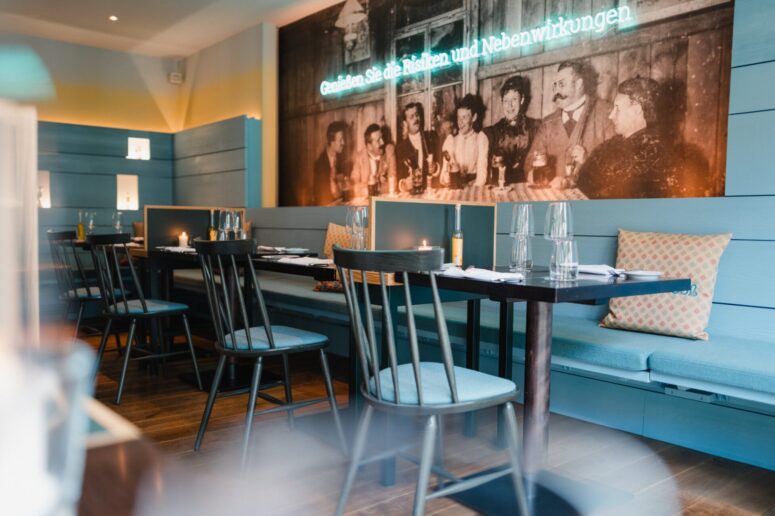 Ein gemütliches Restaurant mit hellblauen Wänden, schwarzen Stühlen, gedeckten Tischen und Sitzbänken mit Kissen. Ein großes Vintage-Foto und eine deutsche Neonschrift schmücken die Wand über dem Sitzbereich.