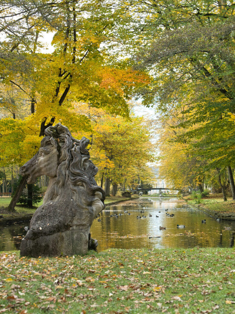 Eine Steinskulptur steht an einem ruhigen Teich in einem Park, der von Bäumen mit gelben und grünen Herbstblättern umgeben ist. Gefallene Blätter bedecken das Gras, und Enten schwimmen im Wasser unter einer kleinen, entfernten Brücke - perfekt für einen entspannten Spaziergang.